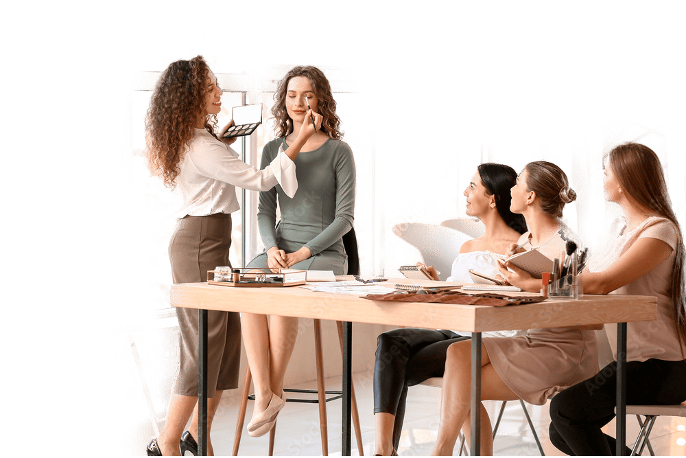 A makeup artist applies makeup to a seated woman's face, demonstrating techniques to an attentive group of four women seated around a wooden table. The table is scattered with makeup products and papers. The setting appears to be a beauty school in Fairfax County, offering cosmetic classes and tutorials.