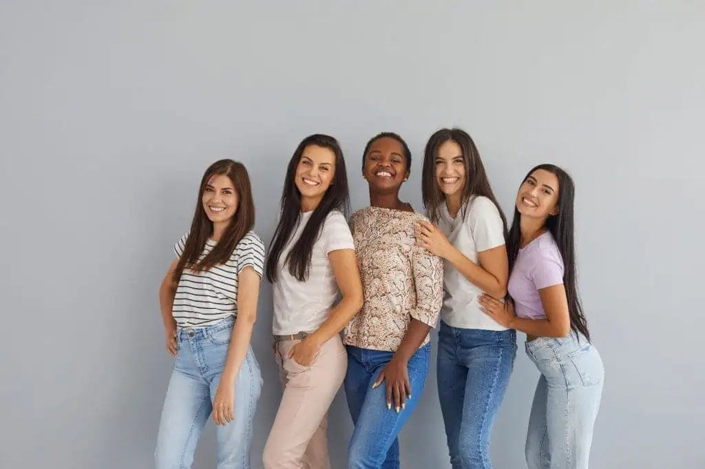 Five women stand closely together against a plain gray background, smiling at the camera. They are casually dressed in jeans and assorted tops, displaying relaxed, cheerful body language that highlights camaraderie and friendships formed during their studies at Beauty School Fairfax County.