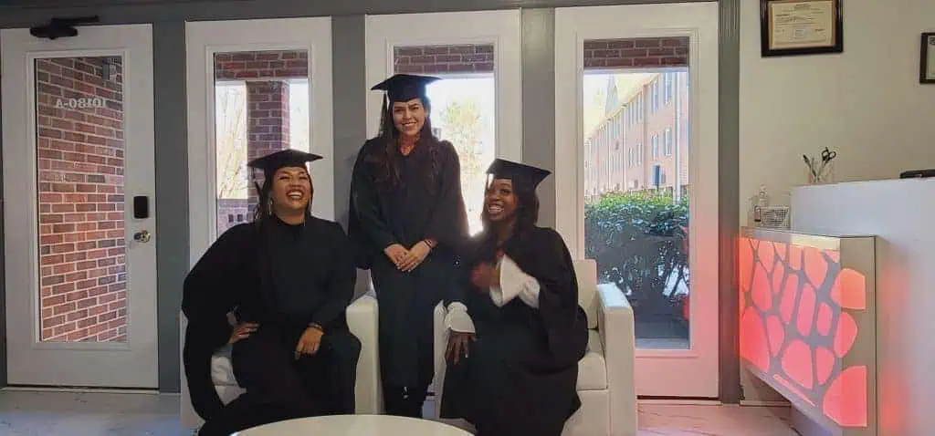 Three graduates in black caps and gowns are in a well-lit room with large windows. Two sit on a white couch while one stands behind them, all smiling and celebrating their accomplishments from Beauty School Fairfax County. A framed certificate hangs on the wall beside the door.