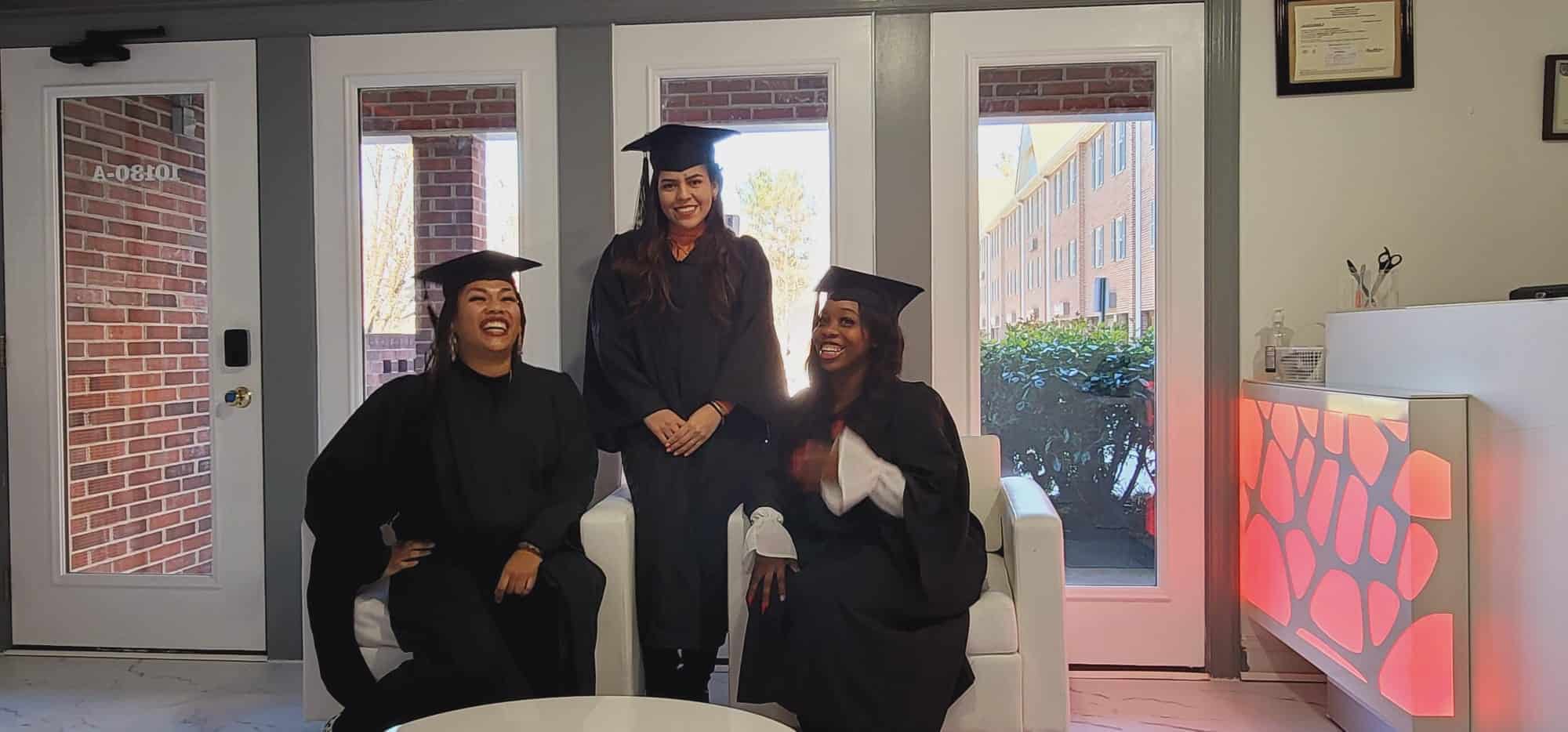 Three graduates in black caps and gowns are in a well-lit room with large windows. Two sit on a white couch while one stands behind them, all smiling and celebrating their accomplishments from Beauty School Fairfax County. A framed certificate hangs on the wall beside the door.