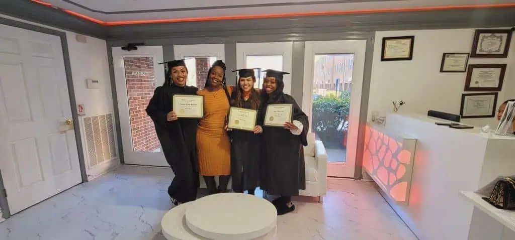 Four smiling individuals in academic regalia hold certificates inside a bright room with modern decor. A woman in a mustard dress stands in the center, celebrating their achievements at the Cosmetology School Fairfax County. The setting includes a white counter, wall certificates, and large windows revealing an outdoor scene.