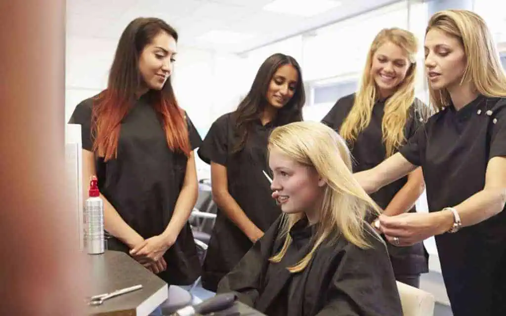 A hairstylist cuts a woman's hair while three other women watch, all wearing black salon capes in a bright, modern salon with large windows.