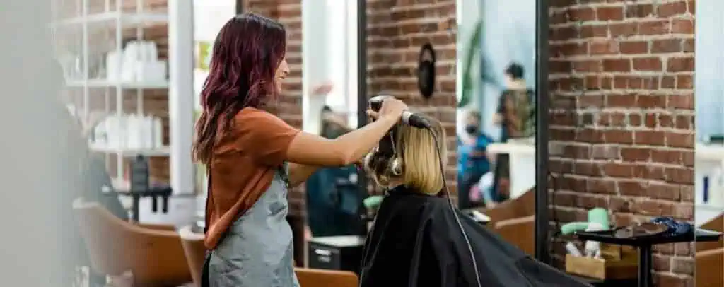 A hairstylist with long, dark hair highlights a client’s hair in a salon. The client wears a black cape while the stylist works, surrounded by brick walls, salon shelves, and other clients and stylists in the background. This scene reflects professional cosmetology training in Fairfax County.