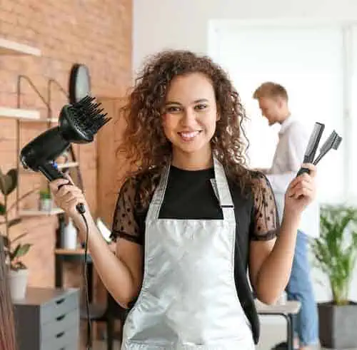 A cheerful woman with curly hair, wearing a silver apron, holds a hairdryer in one hand and combs in the other. She stands in a modern salon with exposed brick walls, showcasing skills she perfected at Beauty School Fairfax County. In the background, a man is seen working diligently.