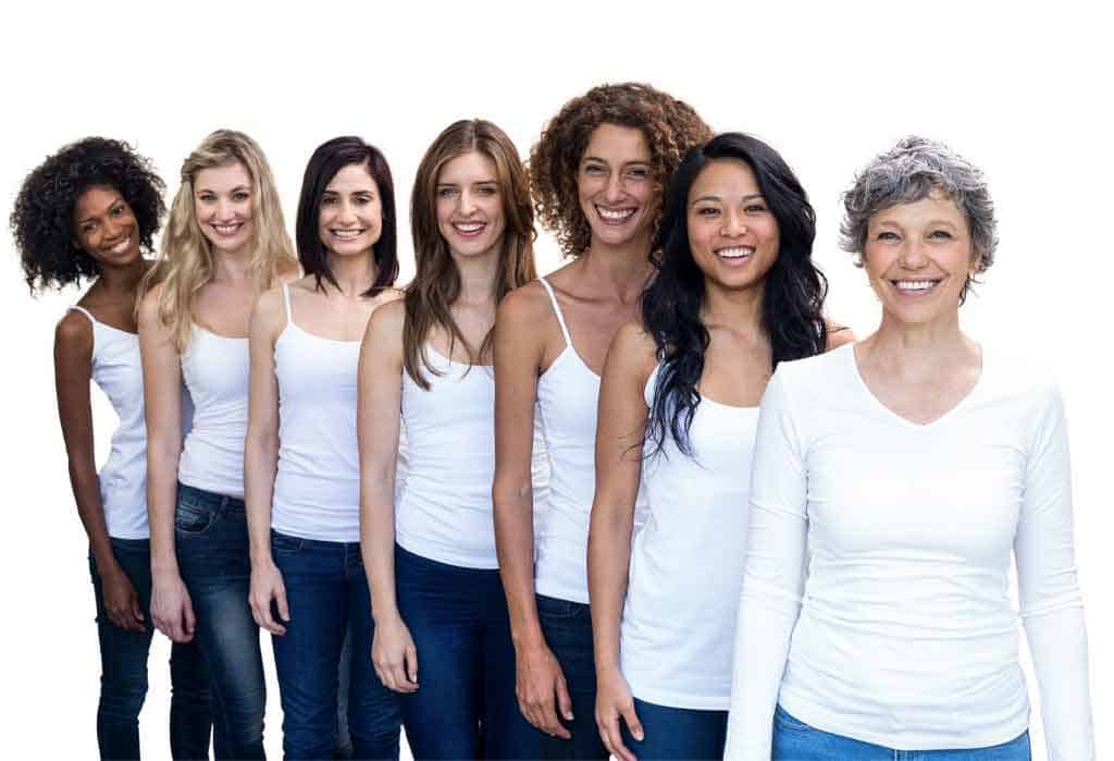 Seven women standing in a row, smiling. They are dressed in white tops and blue jeans and have varied hairstyles and hair colors. The background is plain white, reminiscent of the fresh and professional vibe you'd find at a beauty school in Fairfax County.