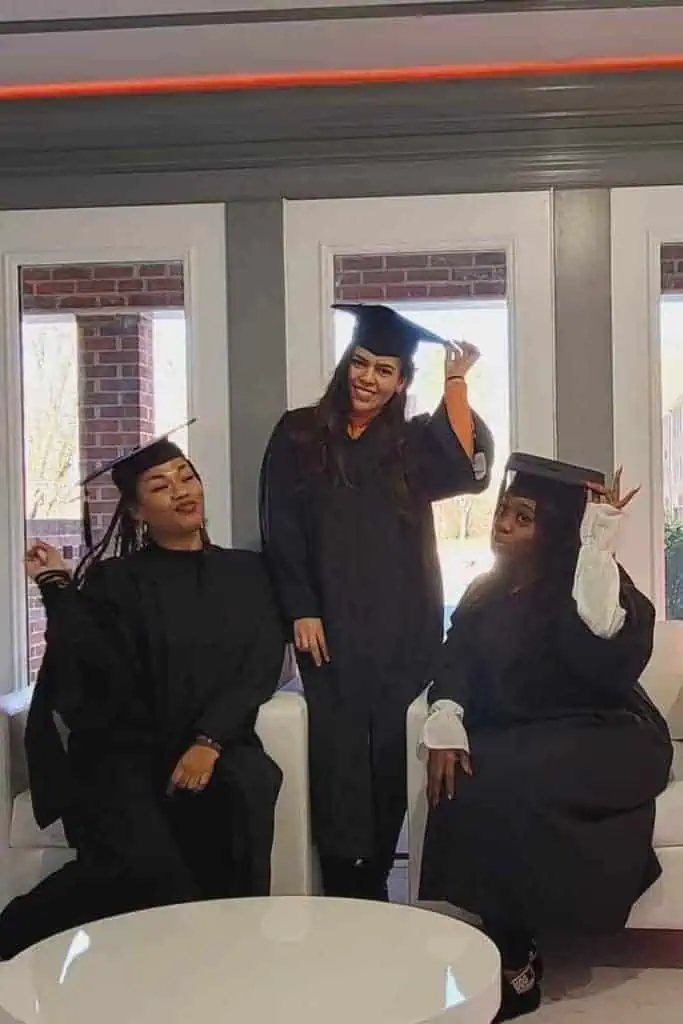 Three graduates from a beauty school in Fairfax County are wearing black caps and gowns, posing inside a room with large windows. The central graduate stands and adjusts their cap, smiling, while the other two are seated, smiling, and making celebratory gestures.