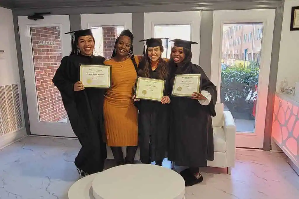 Four people are standing indoors. Three of them are wearing graduation caps and gowns, each holding a certificate from Beauty School Fairfax County. They are smiling at the camera and have their arms around each other. The fourth person is dressed in a yellow dress and standing in the center.