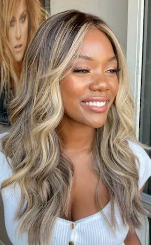 A woman with long, wavy blonde hair and dark roots smiles while looking to the side. She wears a white button-up top. Behind her, a framed photo on the wall displays another blonde woman showcasing lash extensions, highlighting services and training at Beauty School Fairfax County.