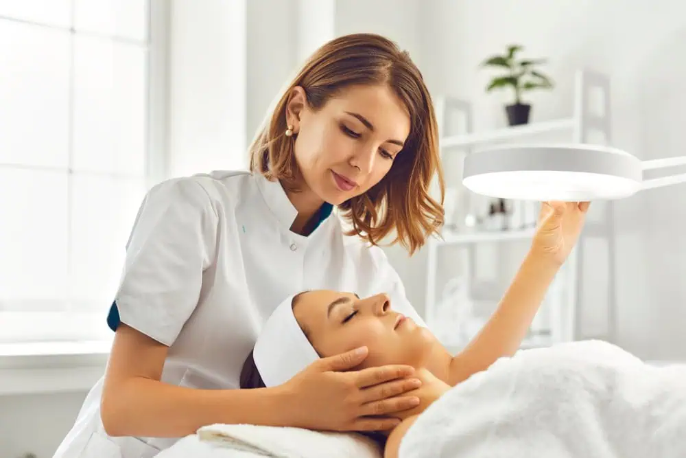 A skincare professional in a white uniform examines a client’s face under a bright magnifying lamp in a spa-like treatment room. The client lies comfortably with a headband securing their hair, eyes closed in relaxation. The space is softly lit and decorated with plants, creating a calming and professional wellness environment.