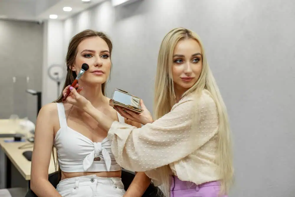 Two women in a room with modern lighting. One with long blonde hair in a polka-dot blouse applies makeup with a brush to the other's face, who is sitting and wearing a white top. They appear focused on the task.