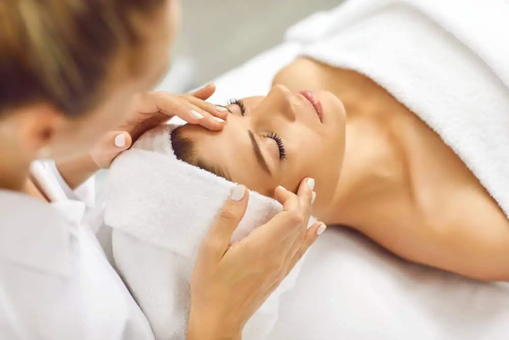 A person receiving a relaxing facial massage in a spa setting. They are lying down with their head wrapped in a towel, eyes closed, while a therapist gently massages their temples with both hands.