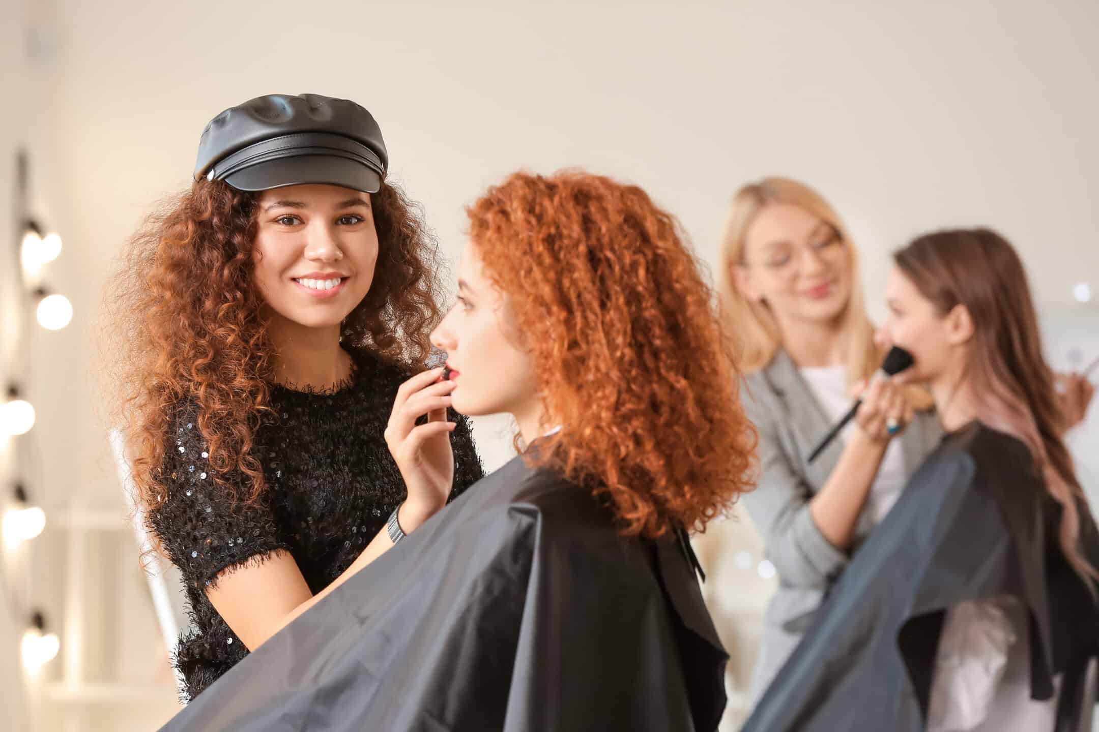 Two women receive makeup in a salon. The first woman with curly hair and a black cap applies lipstick to a seated client with curly red hair. In the background, another stylist applies makeup to a client with straight hair.