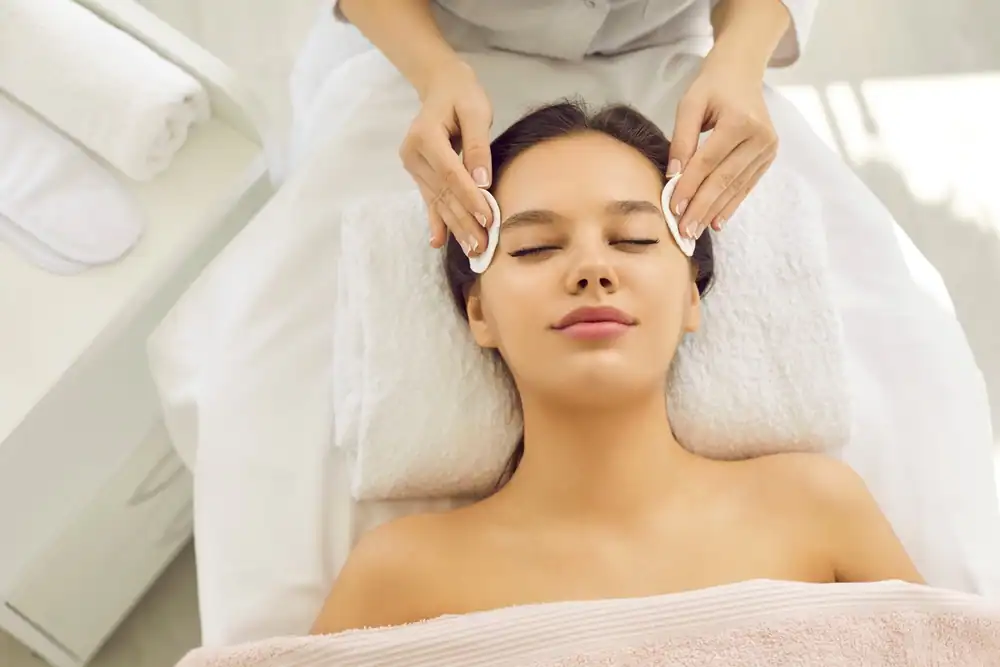 A client receives a relaxing facial treatment at the AVI Career Training spa, where a student esthetician gently applies a skincare mask with professional precision. The client lies on a spa bed with a soft headband keeping her hair back, eyes closed in deep relaxation. The treatment room is clean, well-lit, and designed to create a calming spa atmosphere, reflecting the professional training environment of AVI Career Training.