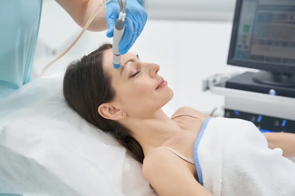 A woman is undergoing a face-lifting procedure in a modern cosmetology clinic. She is lying comfortably on a treatment chair with her eyes closed, while a professional cosmetologist gently uses a specialized device on her skin. The clinical setting is bright, clean, and equipped with advanced skincare technology, emphasizing a professional and safe environment.