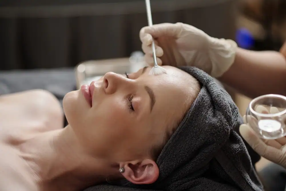 An esthetician wearing protective gloves carefully applies a glycolic acid peel solution with a soft brush to the face of a young woman lying on a spa bed. The client’s eyes are closed, and her head is wrapped in a white towel, creating a serene and professional skincare treatment setting. The atmosphere conveys relaxation, rejuvenation, and advanced facial care.