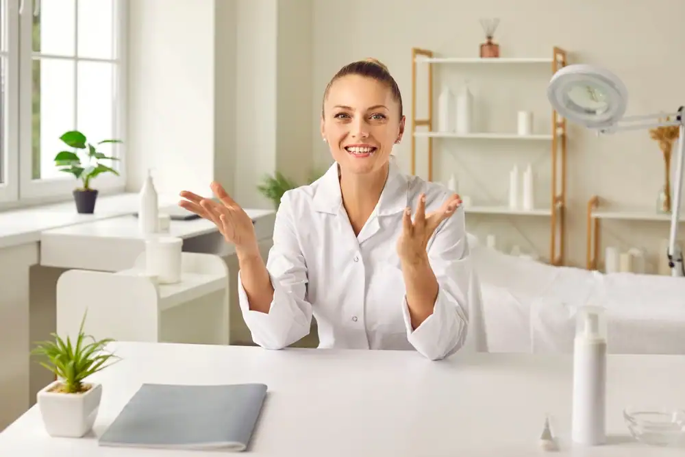 A happy woman cosmetologist and esthetician stands confidently inside her beauty salon, smiling warmly. She appears professional and approachable, representing both her role as a skincare expert and entrepreneur. The salon environment is modern and welcoming, with beauty products and salon equipment neatly arranged, symbolizing her success as a small business owner in the beauty industry.