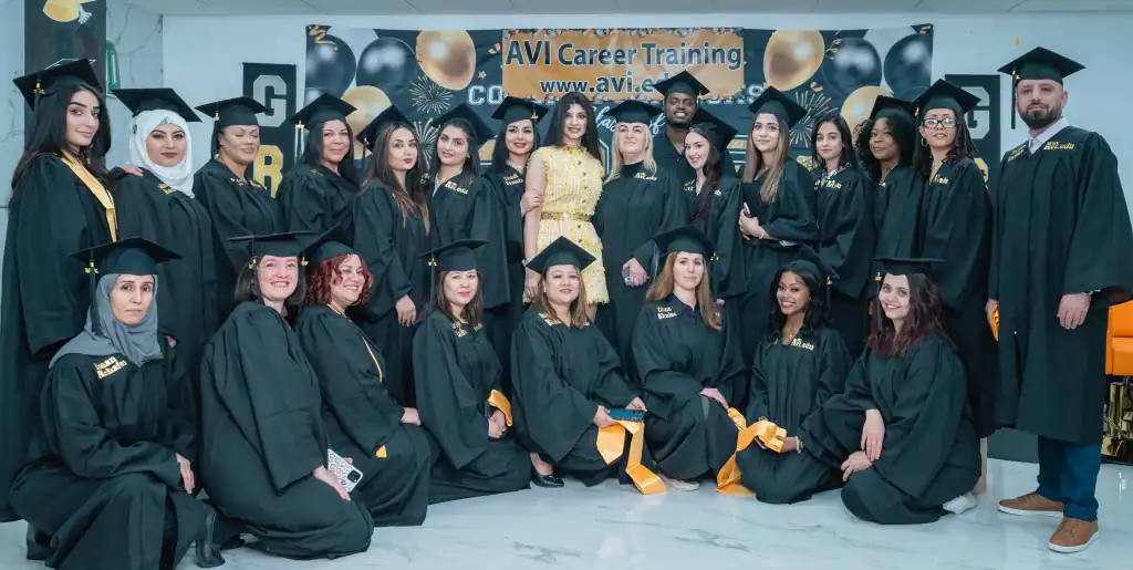 A large group of graduates wearing black caps and gowns poses together indoors in front of a banner that reads "AVI Career Training," celebrating their achievements in cosmetology. Gold and black balloons decorate the background at this VA event.
