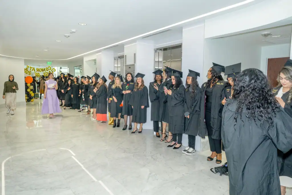 A group of graduates in black caps and gowns, including cosmetology students from Fairfax County, VA, stand in a bright hallway. Some check their phones while a woman in a purple dress stands nearby. Balloons and a “Congrats” sign decorate the background.
