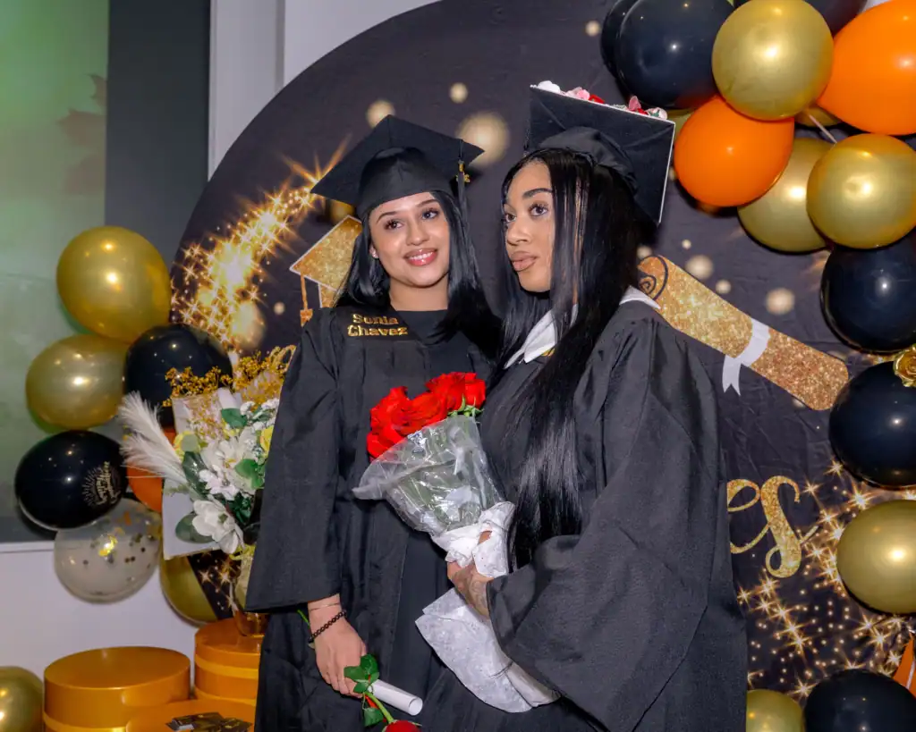 Two graduates in black gowns and caps, celebrating their cosmetology achievements in Fairfax County, VA, smile while holding bouquets and posing in front of a festive balloon backdrop at their graduation celebration.