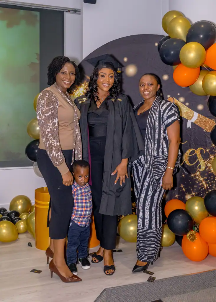 Three women, one in a graduation cap and gown, stand smiling with a young boy at a celebration in Fairfax County, VA, decorated with black, gold, and orange balloons. A "Congrats" sign hints at a special achievement in cosmetology.