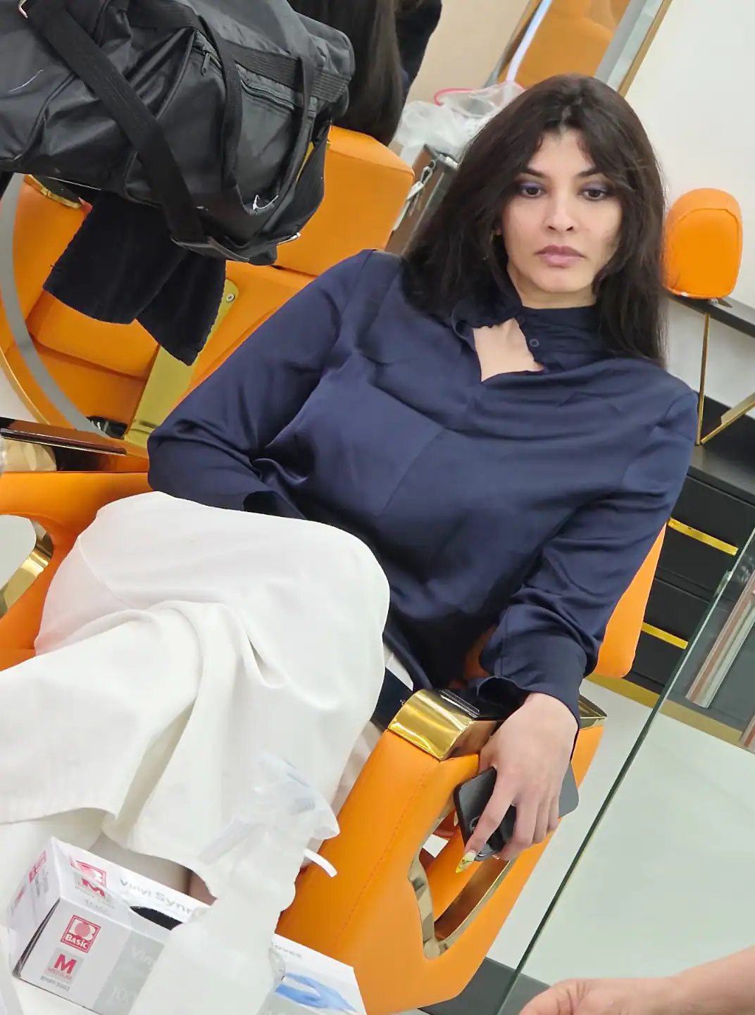 A woman with dark hair, dressed in a navy blouse and white pants, sits thoughtfully on an orange salon chair in a cosmetology Fairfax County, VA studio. A black bag, mirror, and medical supplies are arranged nearby.