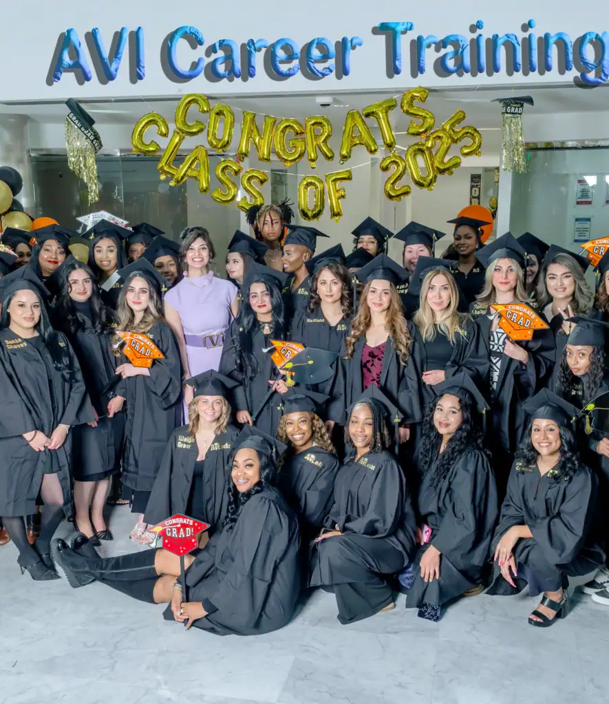 A large group of graduates in black caps and gowns poses indoors under gold balloon letters that read "CONGRATS CLASS OF 2023" at AVI Career Training, VA, celebrating their cosmetology Fairfax County achievements with decorated caps and signs.