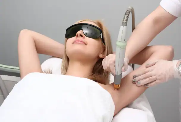 A woman wearing protective glasses receives a laser hair removal treatment on her underarm from a Master Esthetics technician in gloves. She is lying comfortably on a treatment bed covered with a white towel.