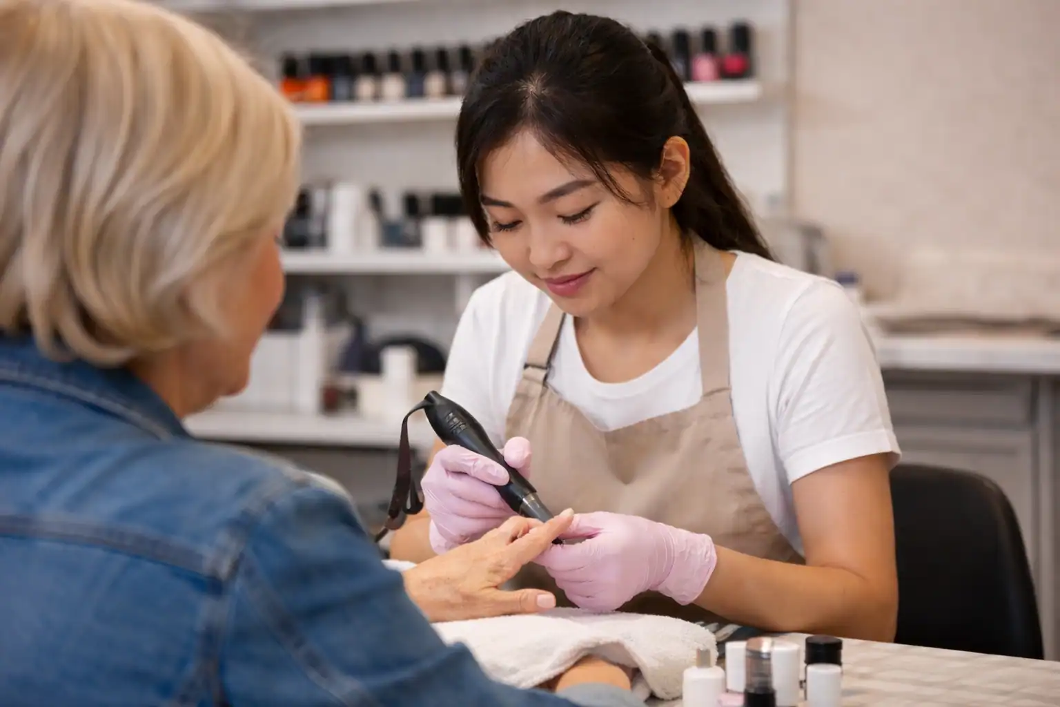 An esthetician in Fairfax County, VA, wearing pink gloves, uses an electric nail file on a customer’s fingernails in a salon, with shelves of nail polish bottles visible in the background.