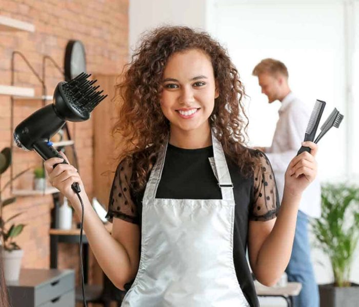 A woman with curly hair, wearing an apron, stands in a salon holding a blow dryer in one hand and two combs in the other. She smiles at the camera while a man is seen in the background conversing. The setting has a modern, brick-walled interior with plants, reminiscent of trendy spots offering Massage Therapy Fairfax County.